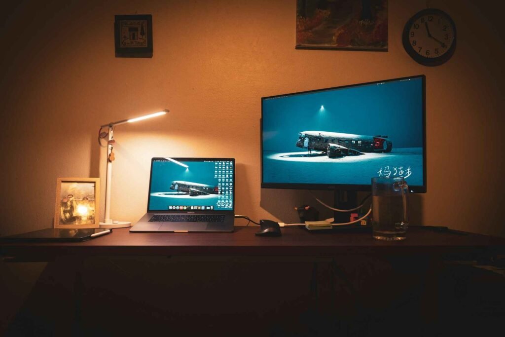 A desk light standing next to a laptop and monitor and lighting the workspace.