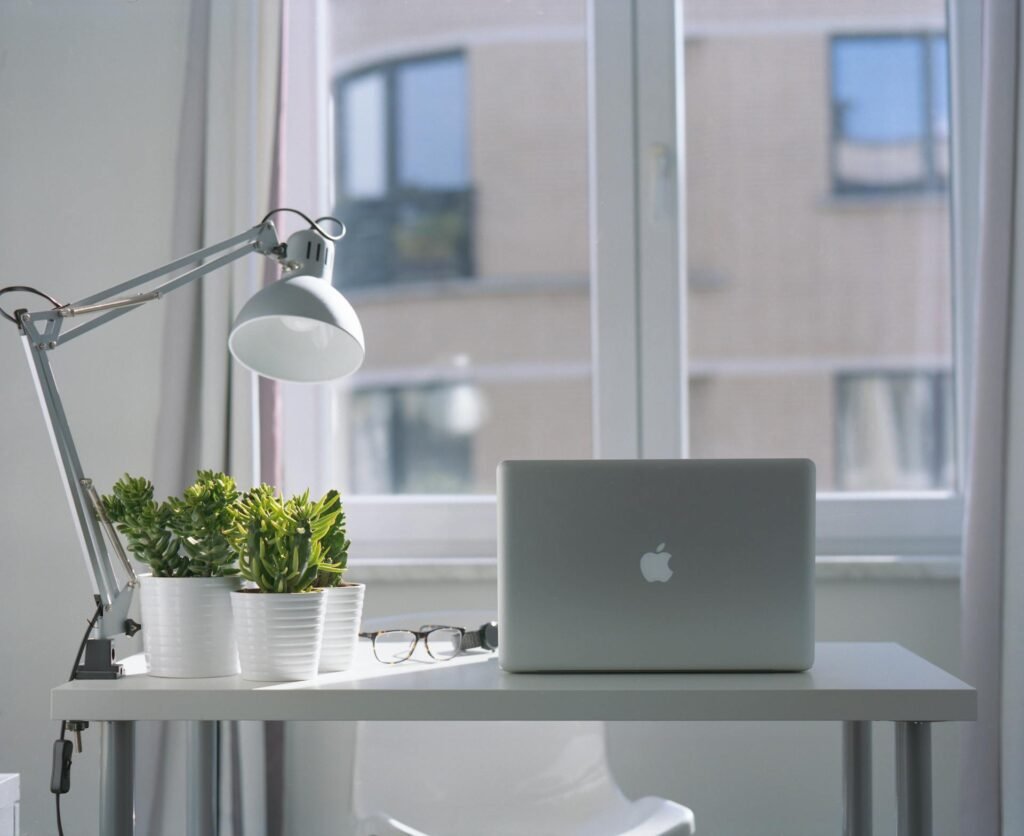 A clip on LED desk light on a white desk with an apple laptop in front of a window.