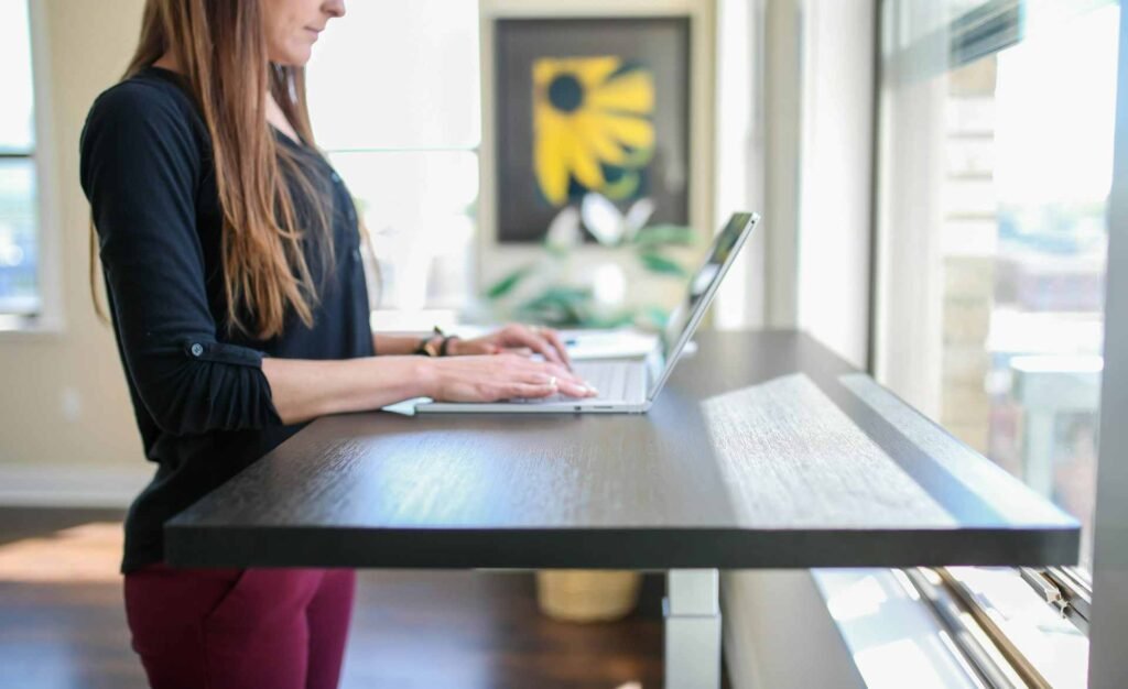 A woman working on a standing desk at a laptop in front of a window.