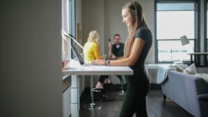A women in grey and black clothing working at a standing desk with good posture. There are 2 people talking in the background.