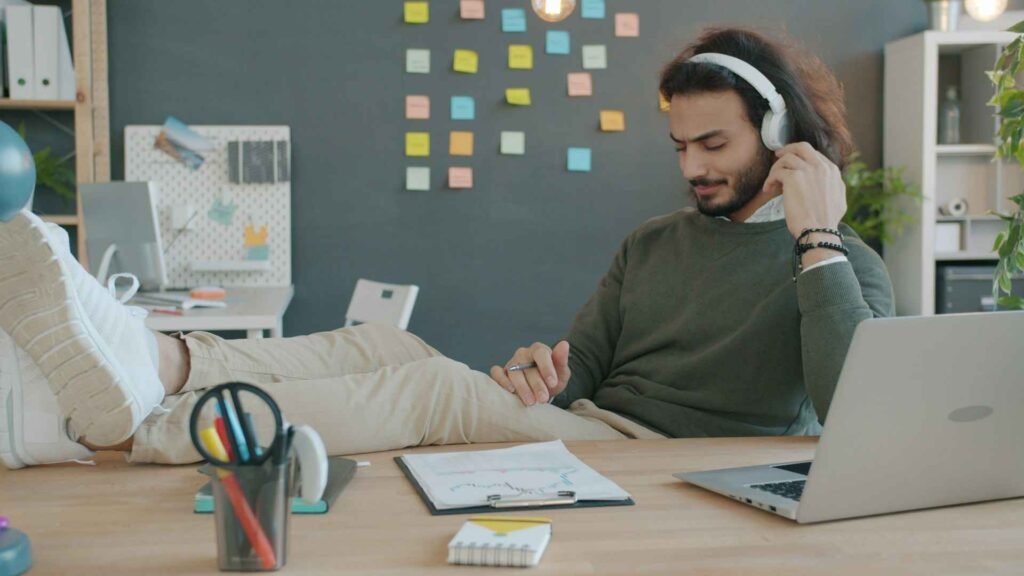 A person with long hair and white headphones listening to music at work. There is a laptop and some notes on a wooden desk in front of him. He also has his legs on the table.
