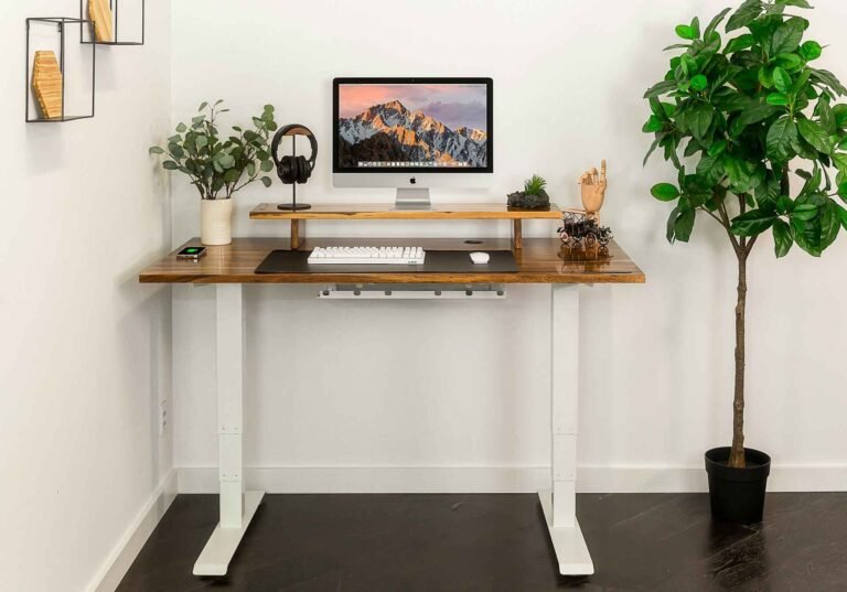 A standing desk with a monitor, headphones, a keyboard, mouse and some plants next to it and on top of it.