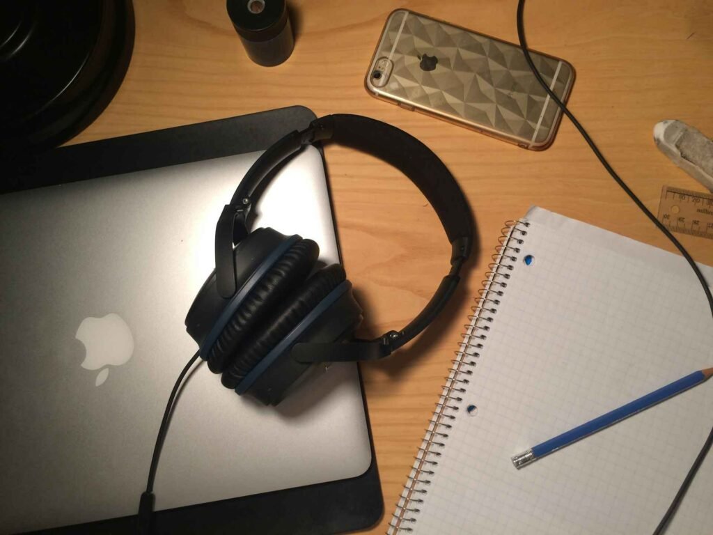 Black corded headphones lying on a wooden table with a laptop and notes.