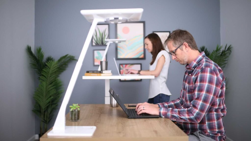 A man sitting at a desk and a women standing at a standing desk in the background. Both are working on laptops with a desk lamp on the desk.
