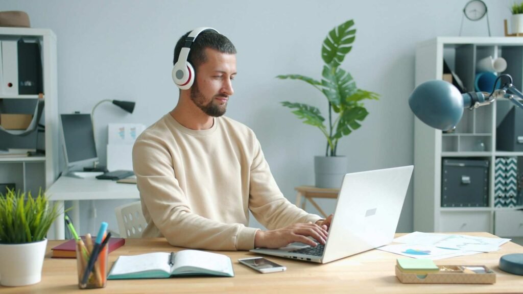 A man with headphones being focused on work on a laptop with some plants around.