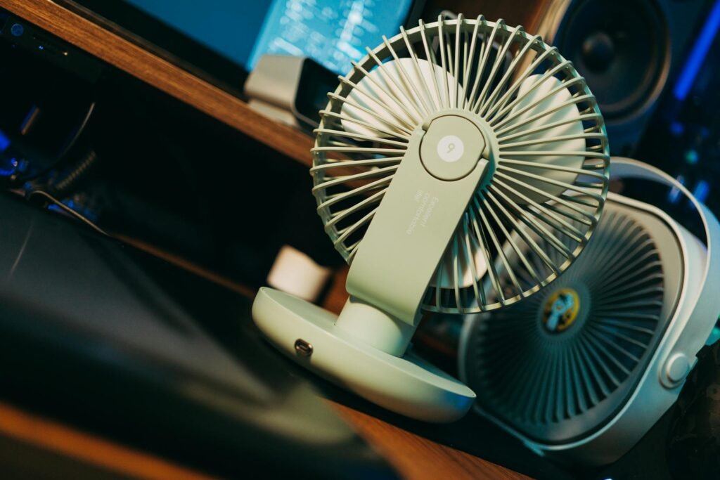 Two desk fans on a wooden desk in front of a monitor.