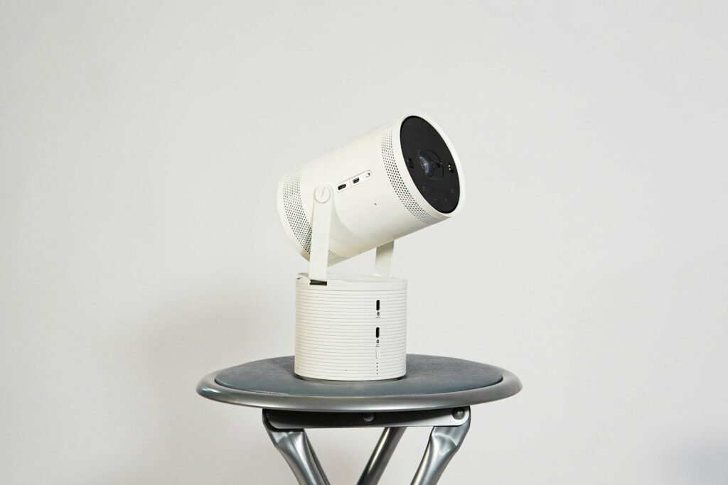A white adjustable mini projector standing on a small grey table in front of a white wall.