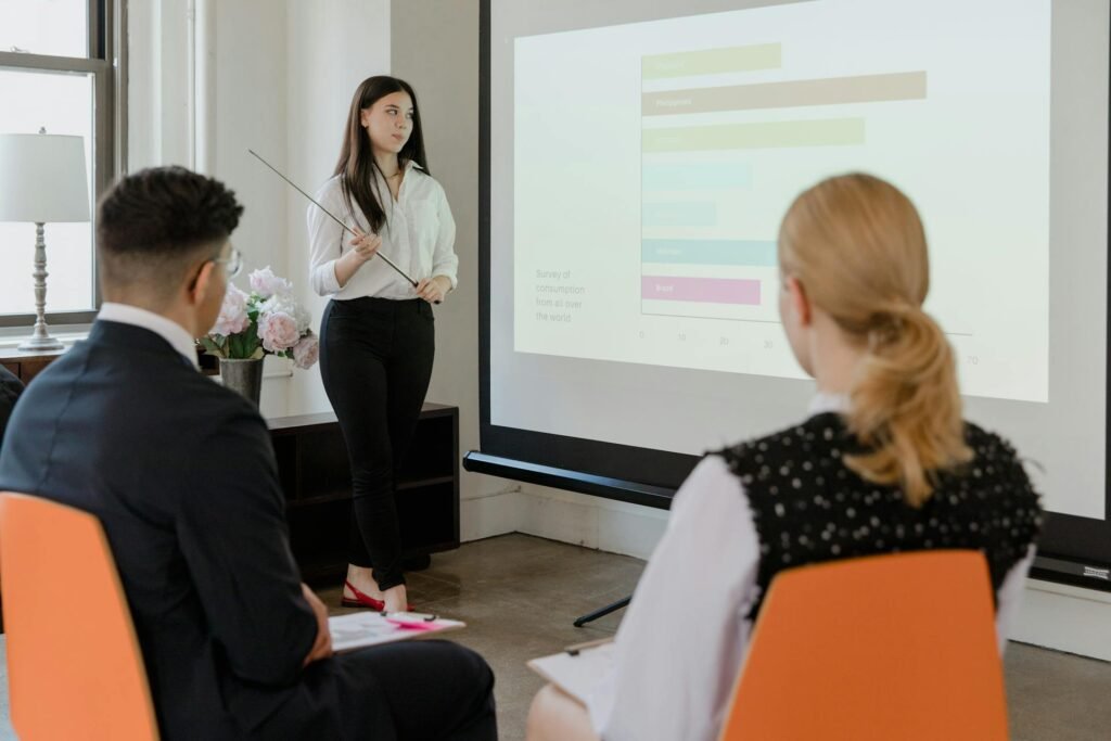 A woman holding a business presentation at work in front of at least two others on orange chairs.