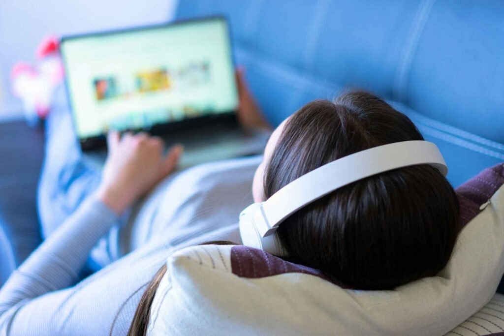 A woman lying on a couch with a laptop and white headphones on.