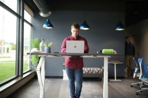 A man in a red shirt working at a standing desk in the middle of the room.