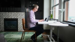 A man working at an adjustable height desk at home in front of a window while sitting.