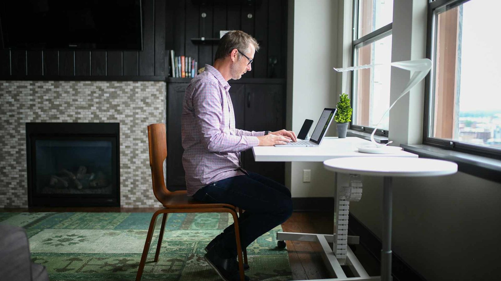 A man working at an adjustable height desk at home in front of a window while sitting.