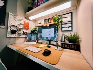 A tiny office space with a monitor, laptop, some plants and a light over it.