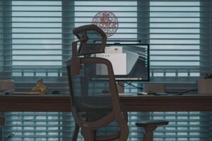 A mesh office chair in front of a desk with a monitor and a window behind it.