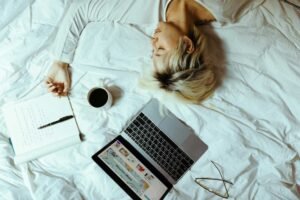 A woman sleeping on a bed with a laptop, some written notes and a cup of coffee.