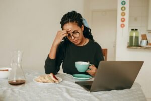 A woman holding her head thinking about something with a laptop and breakfast in front of her.