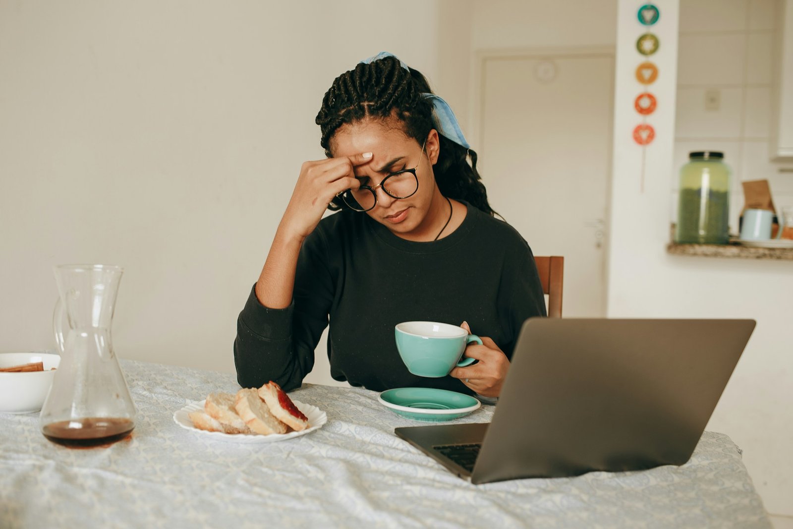 A woman holding her head thinking about something with a laptop and breakfast in front of her.