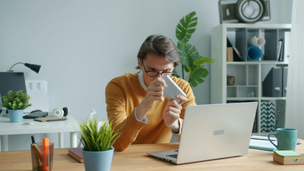 A man with glasses in an orange shirt playing with his phone at work.