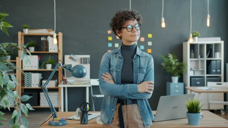 A woman with crossed arms standing in front of a table at work and looking away.