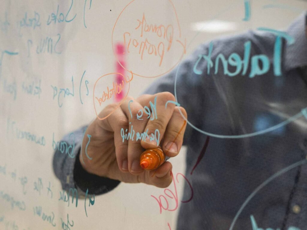 A person writing on a whiteboard with orange and blue markers.